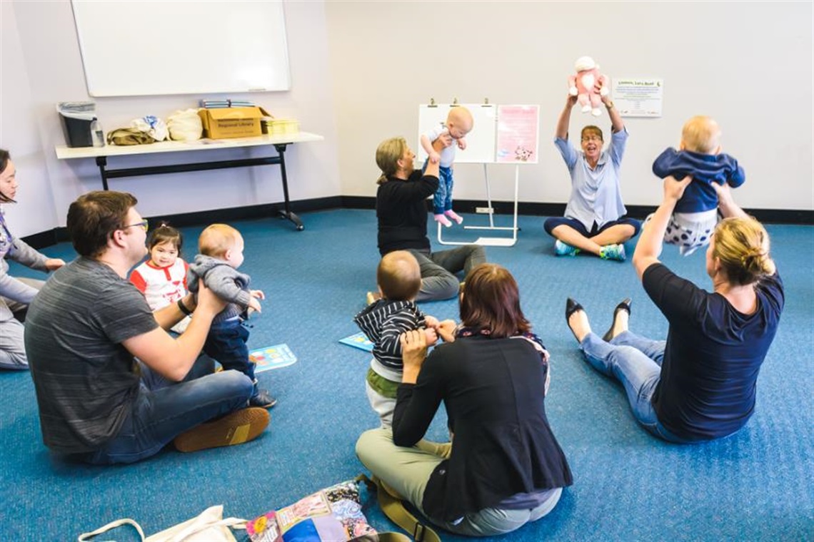 Parents and their bababies sitting on the floor doing an action song with library staff member