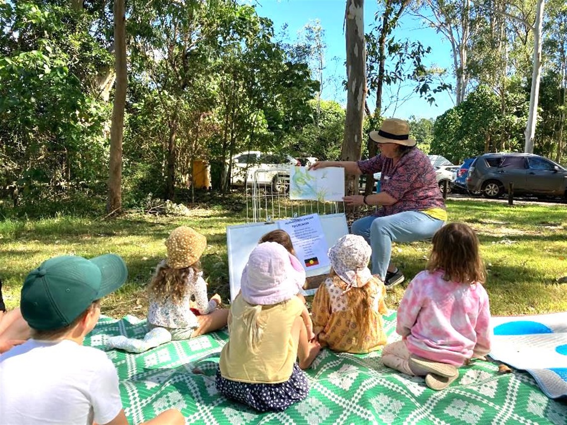 Group of children listening to the story time presenters at the front, all sitting outside in botanical gardens