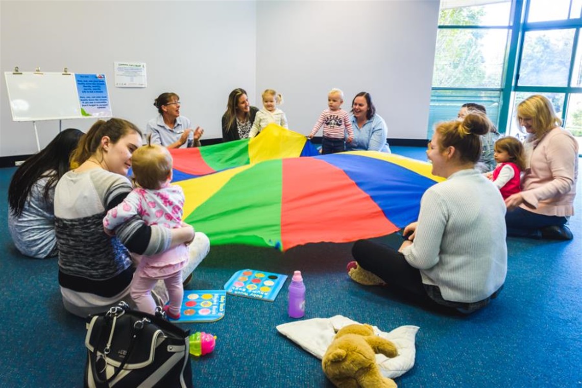 a group of mums and toddlers sitting in a circle with a rainbow parachute