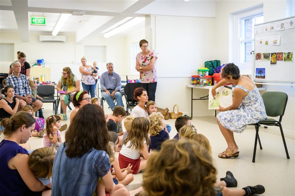 Person sitting at the front of the room on a chair reading to children and their carers sitting on the floor