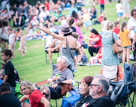 People sitting on folding chairs and picnic rugs at an outdoor event.
