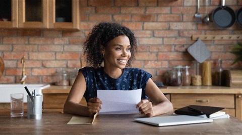 A woman smiles as she holds a piece of paper. She is sitting behind a deck with a laptop and a diary on it.