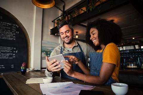 A man and woman, both wearing aprons, behind the front desk at a cafe checking information on a tablet.