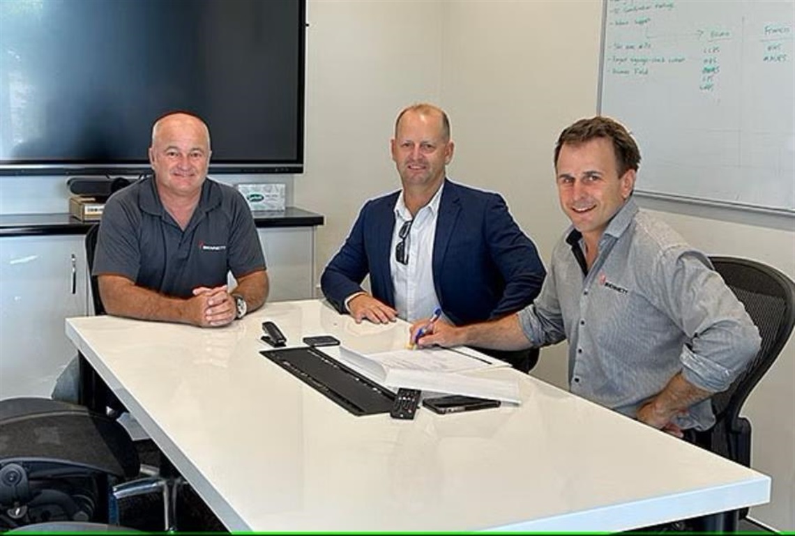 Three men sit around a table which has paperwork on it.
