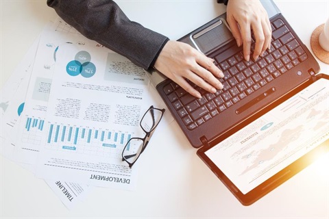 A man types at a laptop with business papers and a pair of glasses on the table next to the laptop.