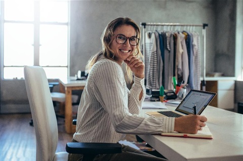 A woman sits at a laptop with a hanging rack full of garments in the background.